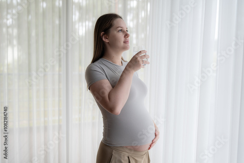 Pensive pregnant gen Z woman drinking water near window. Motherhood, pregnancy, hydration during pregnancy concept photo