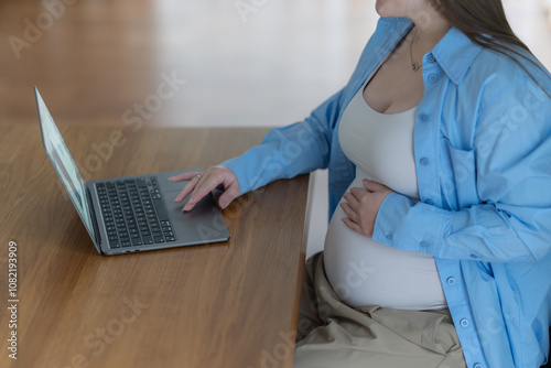 Pregnant business woman using a laptop and holding one hand on her belly while working in the home office.