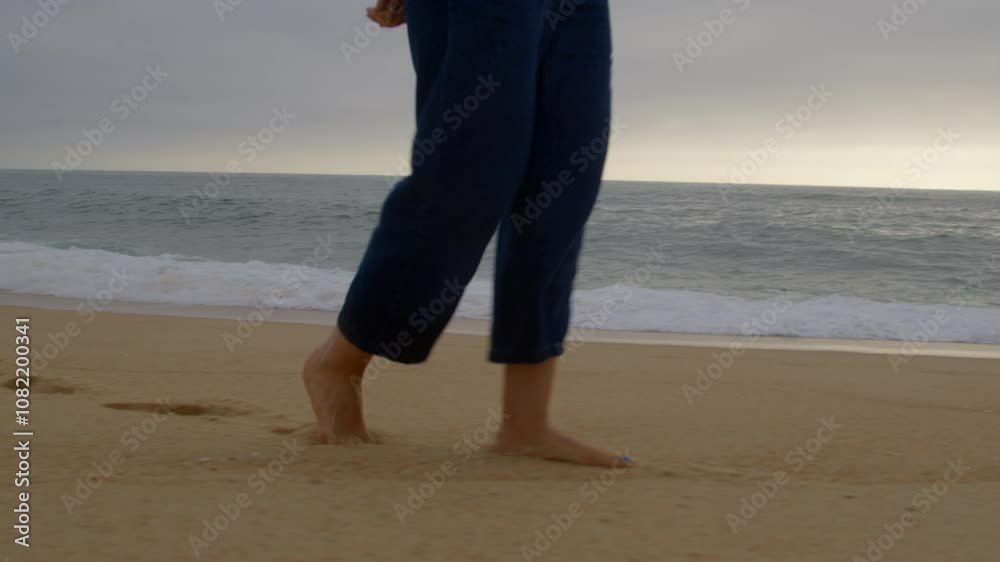 Close-up of a person walking barefoot along a sandy beach at sunset, with ocean waves in the background. The person's face is not visible, only legs and feet captured Close-up of a person walking barefoot along a sandy beach at sunset, with ocean waves in the background. The person's face is not visible, only legs and feet captured