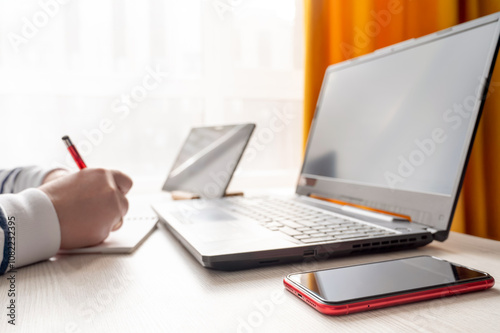 Obraz na płótnie A woman is seated at a desk in front of a laptop, taking notes in a notebook that is lying on the desk. A tablet is also visible on the desk. The background is blurred w salonie
