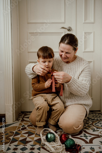 Grandmother and grandson sit on the floor in the kitchen and tie ribbons on Christmas  glass vintage balls.