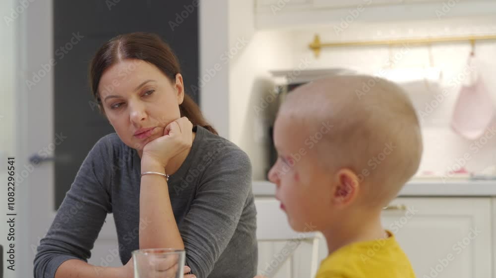 Adult 44 year old woman with tired face watches her child at the kitchen table. Woman smiles tiredly and makes her son eat lunch. Adult 44 year old woman with tired face watches her child at the kitchen table. Woman smiles tiredly and makes her son eat lunch.