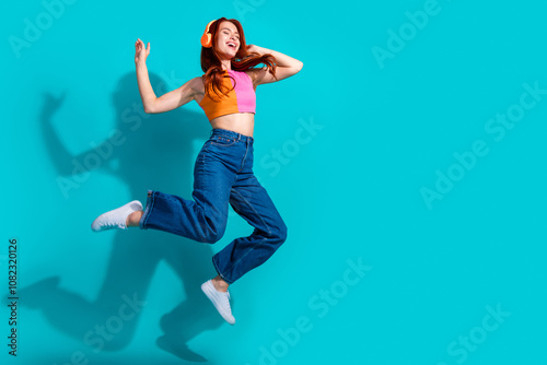 Joyful young redhead woman in a summer casual outfit jumps against a vibrant teal background