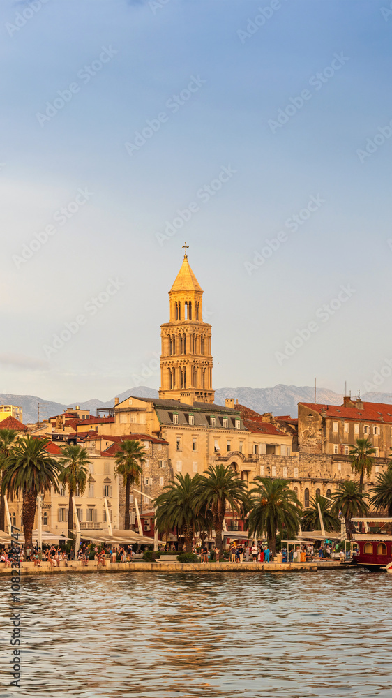 Fototapeta premium Sunset on the lively Riva promenade in Split, Croatia. Lined with palm trees and historic buildings, the waterfront is filled with people and colourful boats.