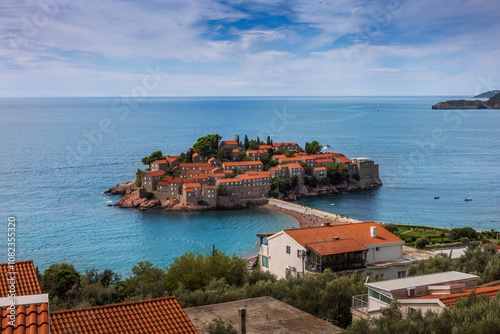 The beautiful island of Sveti Stefan near Budva, Montenegro. Historic red-roofed houses sit on the island in the blue Adriatic Sea, connected to the shore by a narrow isthmus under a partly cloudy sky photo