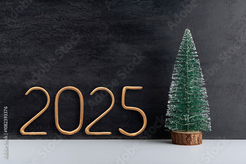 Artificial small Christmas tree on wooden stand and golden number 2025 on black background. New year