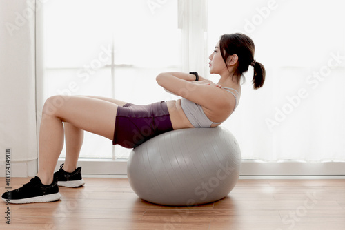 Asian sporty women sitting on fitness ball for doing abs exercises at living room. Joyful beautiful female works out at home. Sporty girl engage in exercises workout. Healthy sport hobby lifestyle.