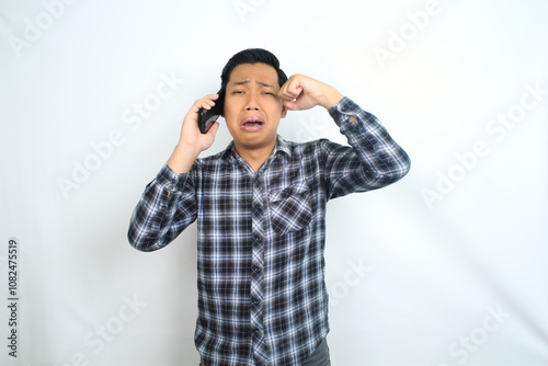 isolated sad asian man crying while talking on cellphone wearing casual plaid shirt