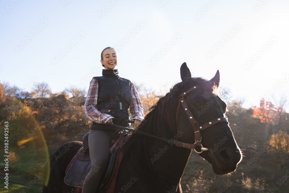 Obraz premium Front view of young cheerful female equestrian riding black horse in countryside in warm sunny autum day, portrait of happy girl on horse, equestrian sport, rider on animal outdoor for adventure
