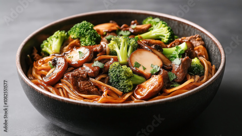 Bowl of stir-fried noodles with broccoli, mushrooms, and sliced beef garnished with fresh herbs in a dark ceramic bowl on a gray background