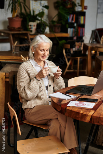 A beautiful mature woman with gray hair savors a drink, immersed in her work at a cafe. photo