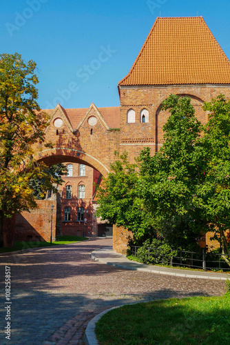 Medieval castle , entrance with main gate and arch with red wirpich. There are walls of the same material all around, the road is paved with old paving stones. There is a lawn and green trees.