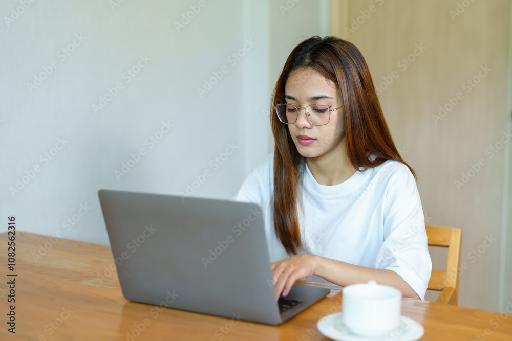 Asian woman with glasses working intently on a laptop at a wooden table, dressed in casual white shirt, with a cup of coffee beside her, focused on her screen in a quiet indoor setting.