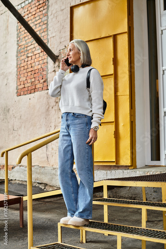 A gray haired woman stands on steps, chatting animatedly on the phone in casual attire.