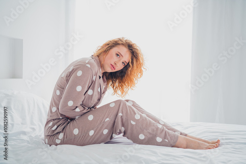 Young woman with red hair in polka dot pajamas sitting on a bed in a bright bedroom photo