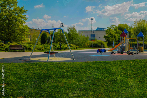 A children's playground in the heart of a city park, surrounded by neatly trimmed trees and a lush green lawn. There’s a white and blue swing set and a bright red slide, with the city skyline in the b