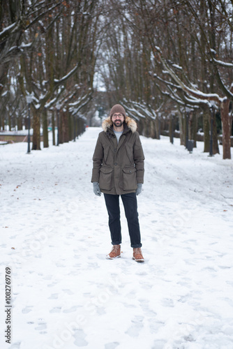 Hombre joven con ropa de invierno (Abrigo, gorro y botas) en jardin nevado con árboles de fondo en Zaragoza (España)