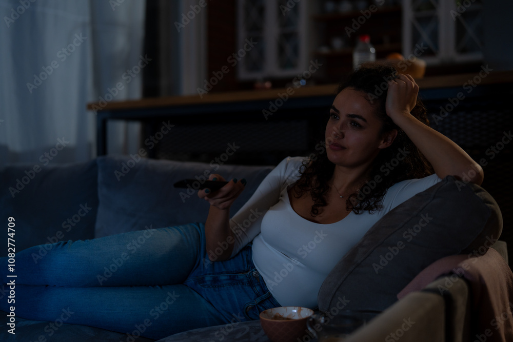 Obraz premium In the evening, a happy curly-haired woman sits comfortably on the couch, enjoying television in a living room lit solely by the TV's glow.