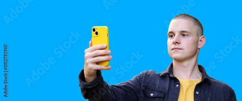 Young man taking a selfie with a yellow phone against a bright blue background in a casual setting