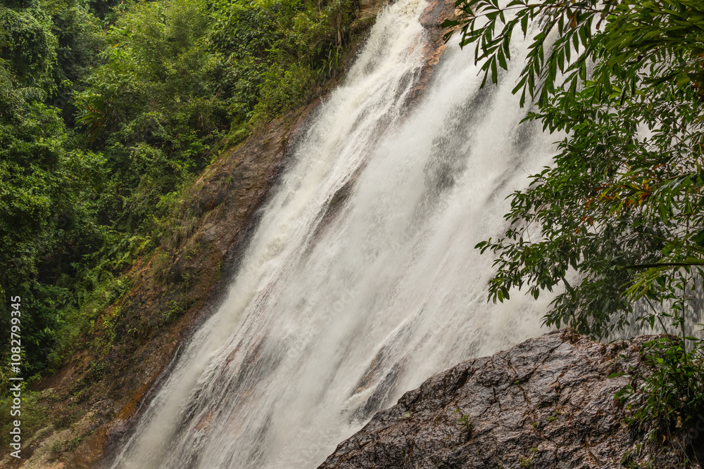 A powerful waterfall tumbling down from a steep rocky cliff surrounded by green foliage. The water falls down with force, creating fog and a sense of movement. A powerful waterfall tumbling down from a steep rocky cliff surrounded by green foliage. The water falls down with force, creating fog and a sense of movement.