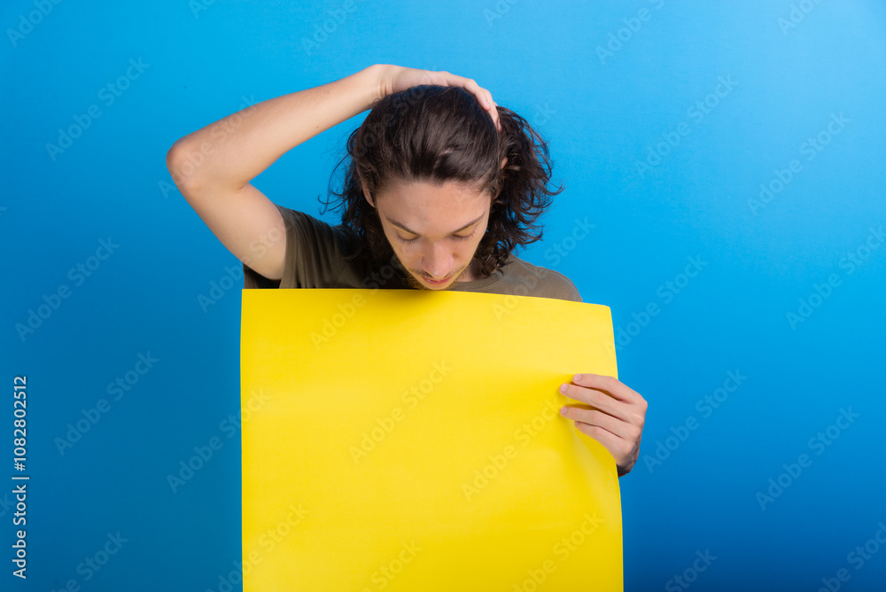 young black haired man holding an empty yellow colored poster isolated on blue background. young black haired man holding an empty yellow colored poster isolated on blue background.