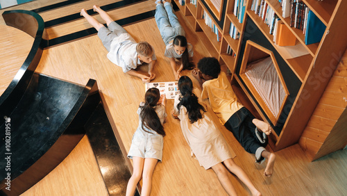 Group of diverse children lying down in circle while reading a book at library. Top view of girl sharing a magazine while pointing at interested topic and talking with lovely friends. Edification. photo