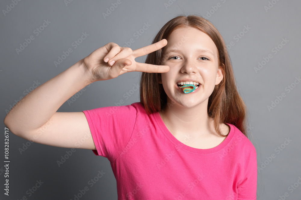 Teenage girl eating tasty rainbow sour belt while showing v-sign on grey background Teenage girl eating tasty rainbow sour belt while showing v-sign on grey background