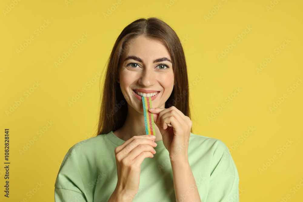 Young woman eating tasty rainbow sour belt on yellow background Young woman eating tasty rainbow sour belt on yellow background