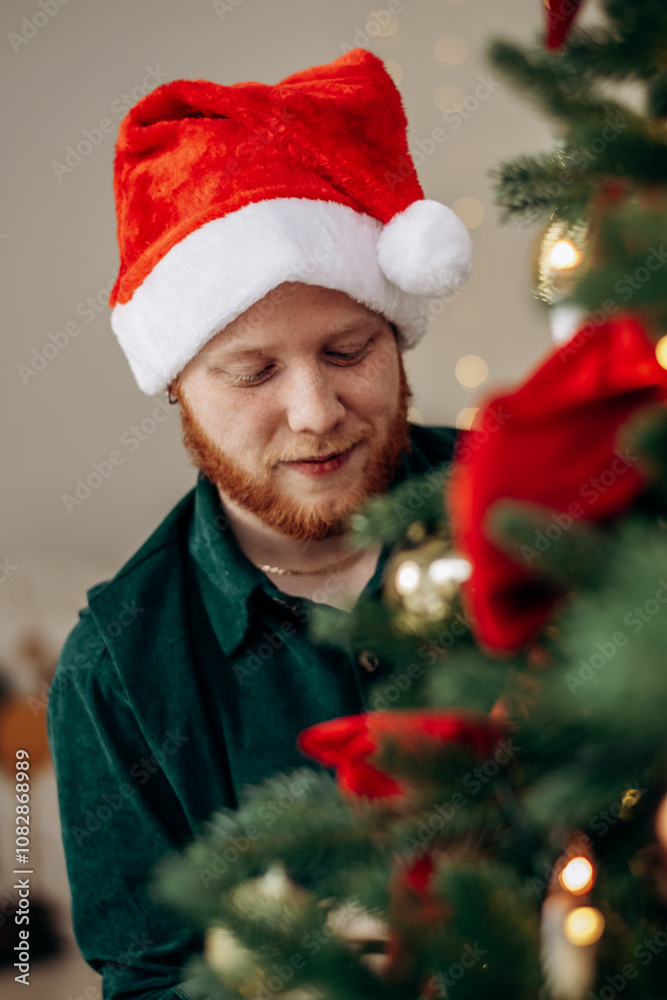 A young red-haired bearded man in the Santa Claus hat is decorating a Christmas tree at home.A New Year's . and Christmas concept. A young red-haired bearded man in the Santa Claus hat is decorating a Christmas tree at home.A New Year's . and Christmas concept.