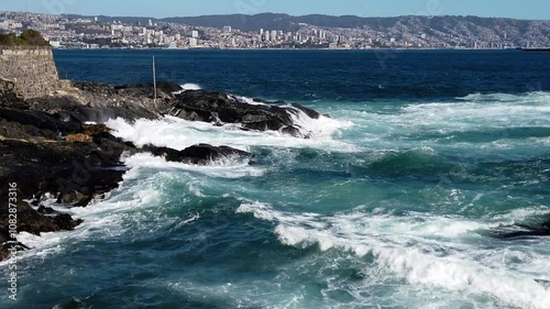 Ocean waves crash against a rocky coast, showcasing the vibrant city of valparaiso in the distance under a sunny sky