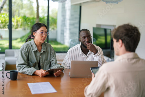 Three young professionals, diverse in ethnicity, are gathered in a modern office. They collaborate using tablets and laptops, dressed in casual business attire, focused on developing innovative ideas.