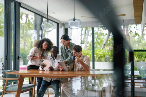 A group of young adult professionals of diverse ethnicities work diligently in a sleek modern office, exchanging ideas over digital devices, reflecting teamwork and strategy.