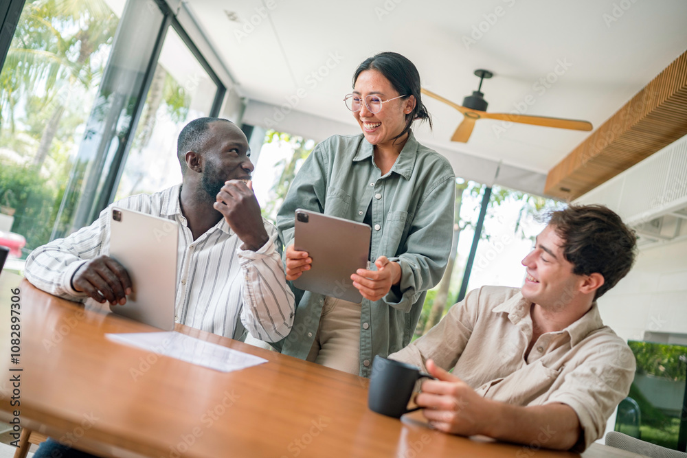 A diverse group of young adults collaborates in a bright office, with a woman leading the discussion using a digital tablet, while two men focus on a laptop and share input.
