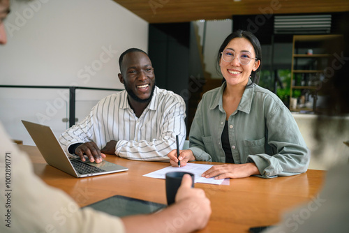 A young Black male and Asian female executive work together, engaging with a laptop in a well-lit office, dressed in professional attire.