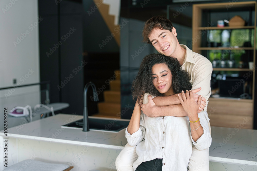 A cheerful young African American woman and her Caucasian partner embrace in a cozy kitchen, reflecting joy and unity in their casual outfits. A cheerful young African American woman and her Caucasian partner embrace in a cozy kitchen, reflecting joy and unity in their casual outfits.