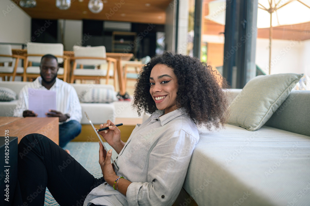 A cheerful young African American businesswoman works on a digital tablet, comfortably seated in a sleek office lounge, with a colleague in the background. A cheerful young African American businesswoman works on a digital tablet, comfortably seated in a sleek office lounge, with a colleague in the background.