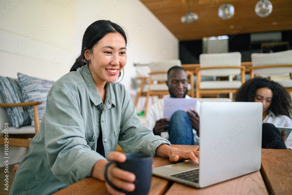 Obraz premium A joyful Asian female young adult uses a laptop in a well-lit home office, her attire casual. A diverse group of colleagues or friends accompany her in the background.