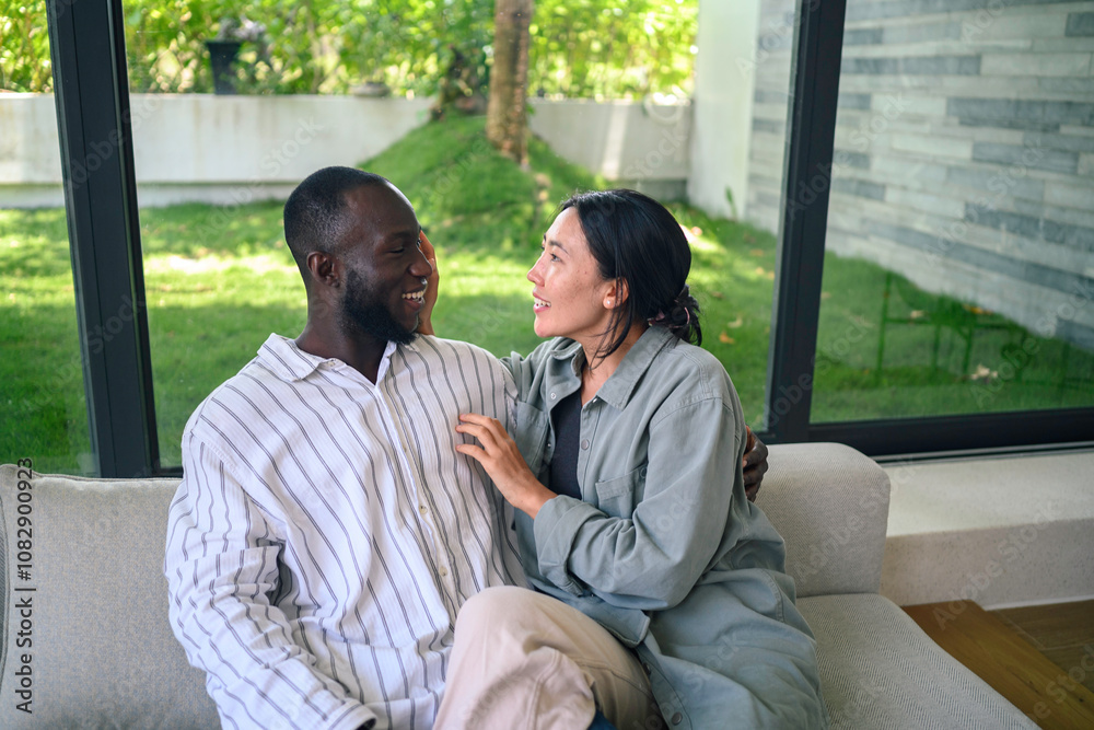 A cheerful Black man and an Asian woman share a relaxed, engaging conversation in their cozy living room, reflecting their close bond and joyful quality time together.