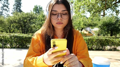 Young brunette woman in glasses and orange shirt listening to music in big white headphones. Woman looking at mobile phone sitting at table against nature background