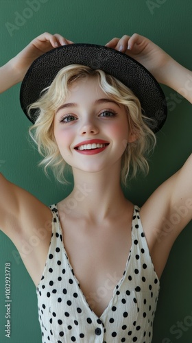 Young woman with blonde hair and a hat smiles joyfully against a plain green ...