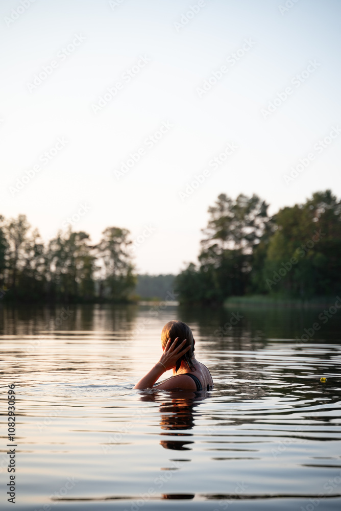 Fototapeta premium A woman swims in a calm lake at sunset, surrounded by reflections of trees and the soft colors of twilight. This peaceful scene highlights relaxation and connection with nature.