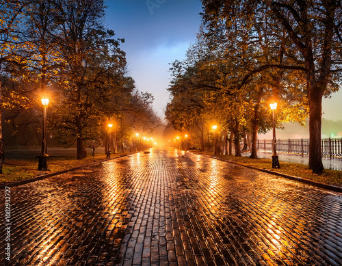 A rain-soaked cobblestone path lined with illuminated lampposts and autumn trees reflects golden light during twilight or evening hours.