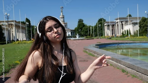 Young brunette woman with white big headphones listening to music dancing in motion against the background of large beautiful buildings. Portrait of a beautiful woman