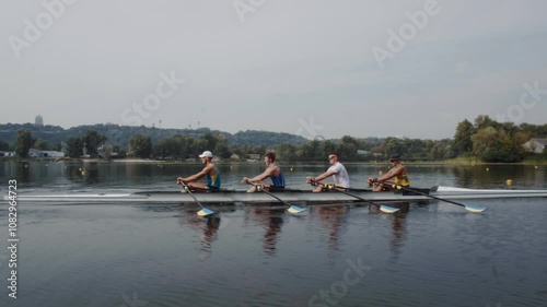 Rowing team training. Side view of 4 young caucasian male rowers, during a rowing practice, athlete sitting in a boat in the river Dnipro, rows through a calm water in autumn. 4k footage. City area in