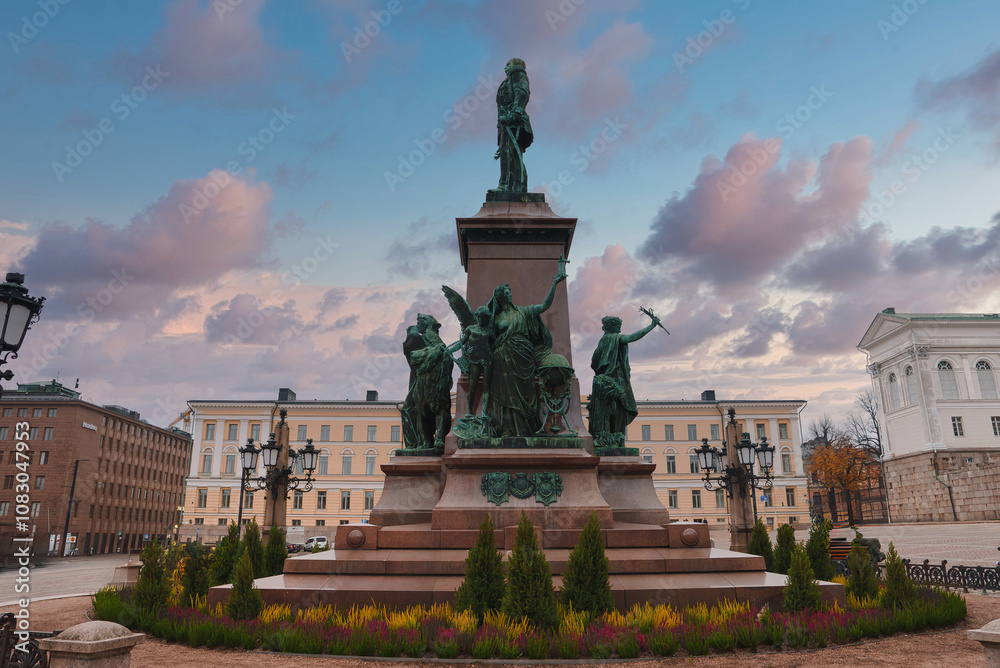 Naklejka premium The statue of Alexander II stands in Senate Square, Helsinki, with the Helsinki Cathedral partially visible. A vibrant sky with clouds forms the backdrop.