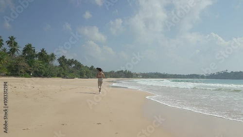 Aerial shot of a woman running along the beautiful Talalla beach in Sri Lanka. The calm ocean waves and tropical scenery set the scene for fitness, relaxation, and exotic travel footage. Ideal for photo