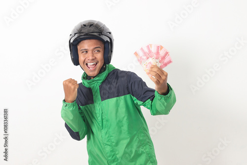 Portrait of Asian online taxi driver wearing green standing against white background, holding a bunch of rupiah money while raising fist, celebrating success photo