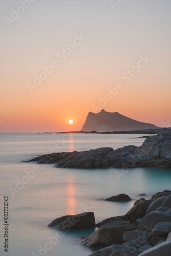 A rocky beach at sunset with the sun setting over the horizon and a silhouette of a headland in the distance.