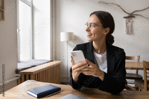 Happy businesswoman sits at desk holding smartphone looking out window with smile, receiving work-related positive notification, taking moment to relax, listen voice audio message. Modern technology photo