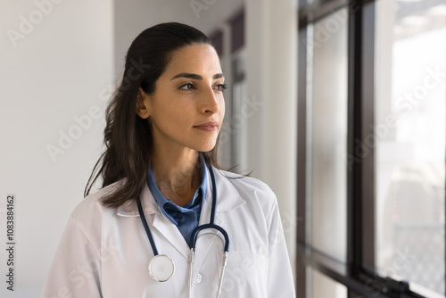 Fotografi Thoughtful Brazilian female doctor in white coat with stethoscope gazing away with serious expression, thinking on medical specialist career, looking out window, deep in thoughts